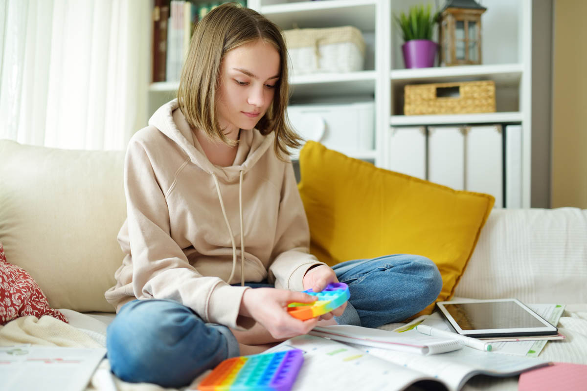 Teenage girl playing with rainbow pop-it fidget toy while studying at home. Teen kid with trendy stress and anxiety relief fidgeting game. Popping the dimples of sensory silicone toy.