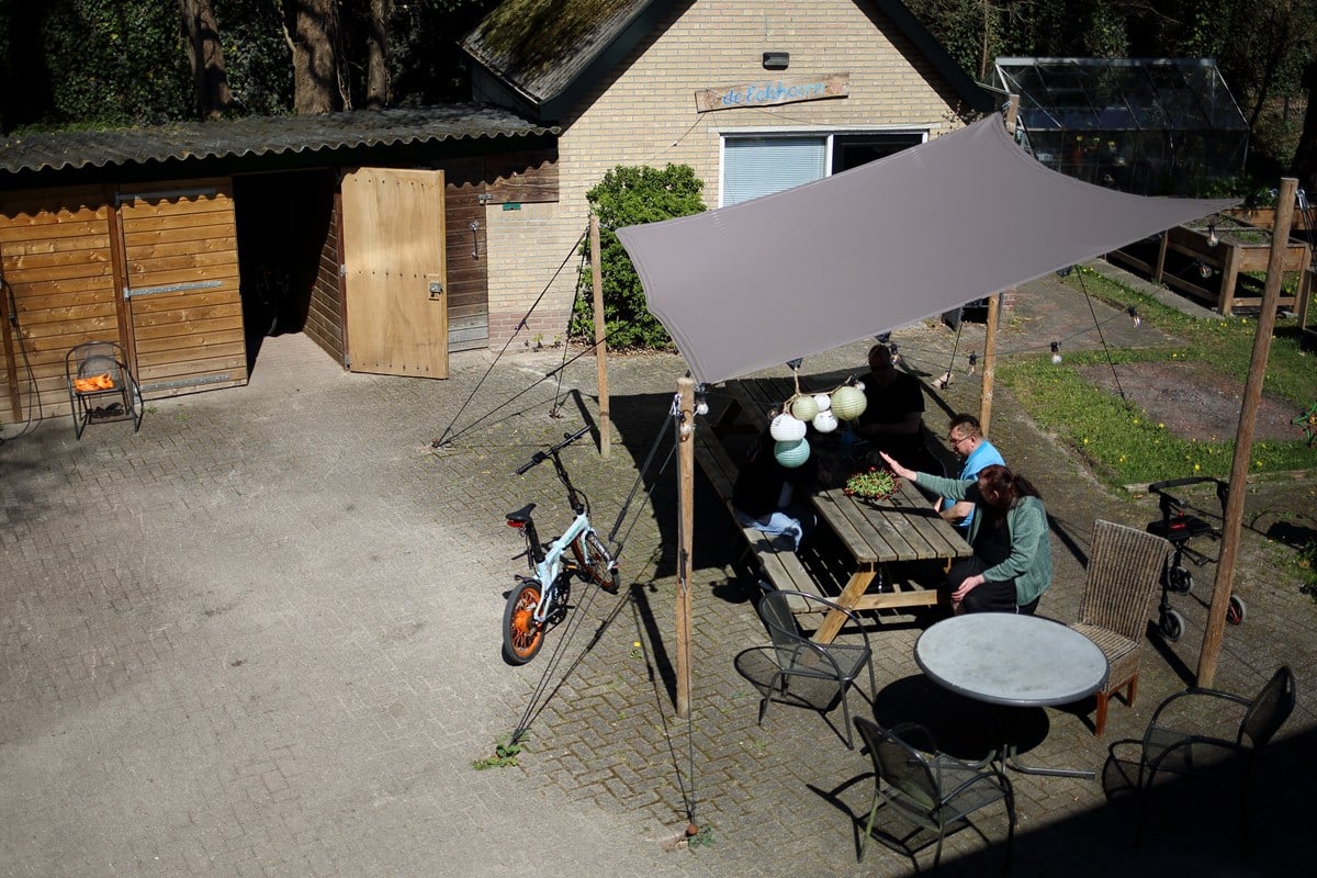 Gezellig buitenterras met houten tafels onder een schaduwdoek, waar mensen samen aan tafel zitten bij een gebouw met de naam “De Eikboom”.