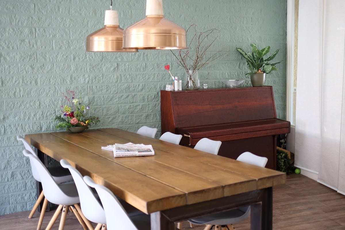 Sfeervolle eetkamer met houten tafel, witte stoelen, groene muur, hanglampen van koperkleurig metaal en een piano tegen de wand.
