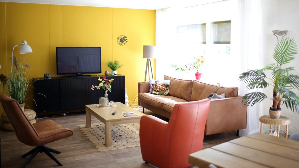 Gezellige huiskamer met gele muur, bruine leren bank, oranje fauteuil en houten salontafel, verlicht door een groot raam met witte gordijnen.