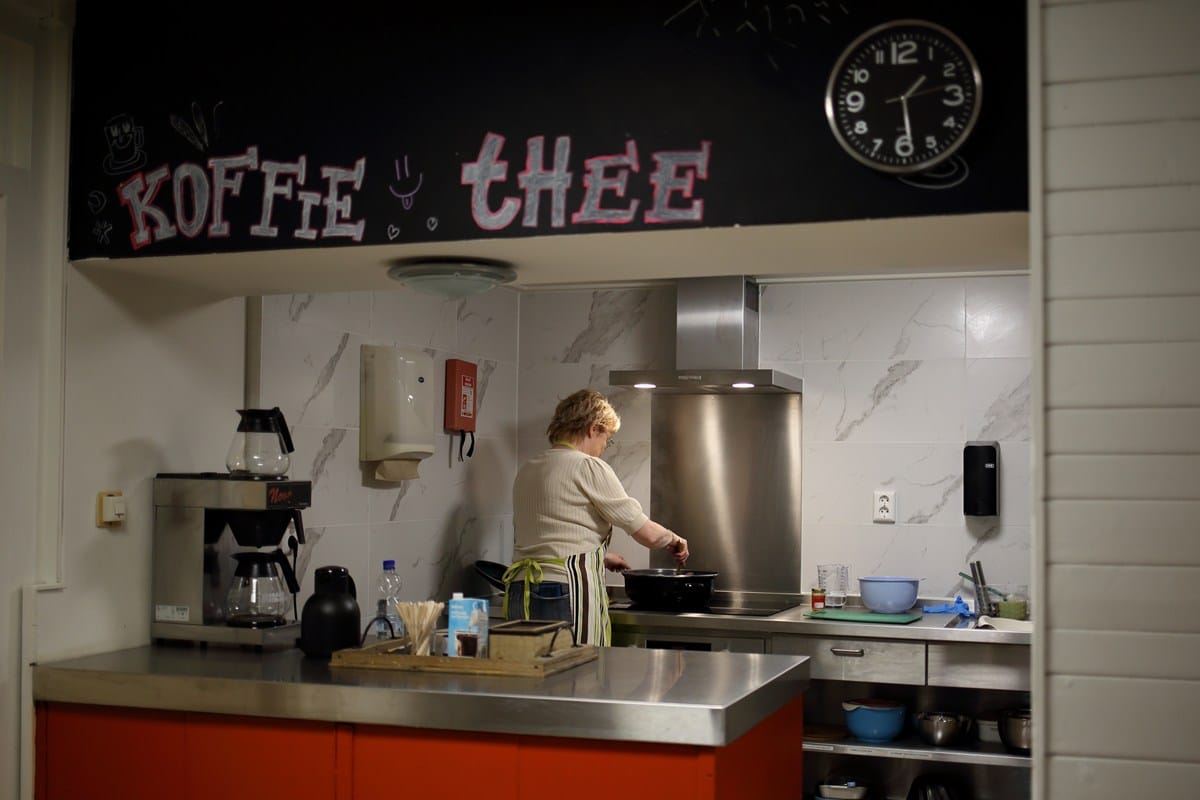 Keuken met een roestvrijstalen aanrecht waar een vrouw staat te koken, met erboven een bord met de woorden "Koffie" en "Thee".