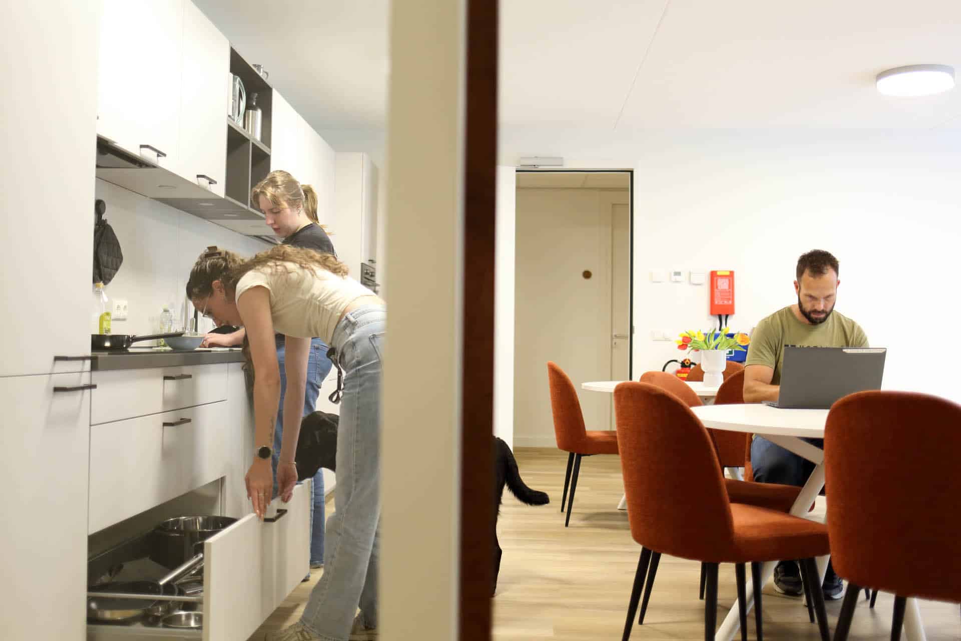 Twee personen bereiden samen eten in een gezamenlijke keukenruimte, terwijl een derde persoon aan tafel werkt op een laptop.