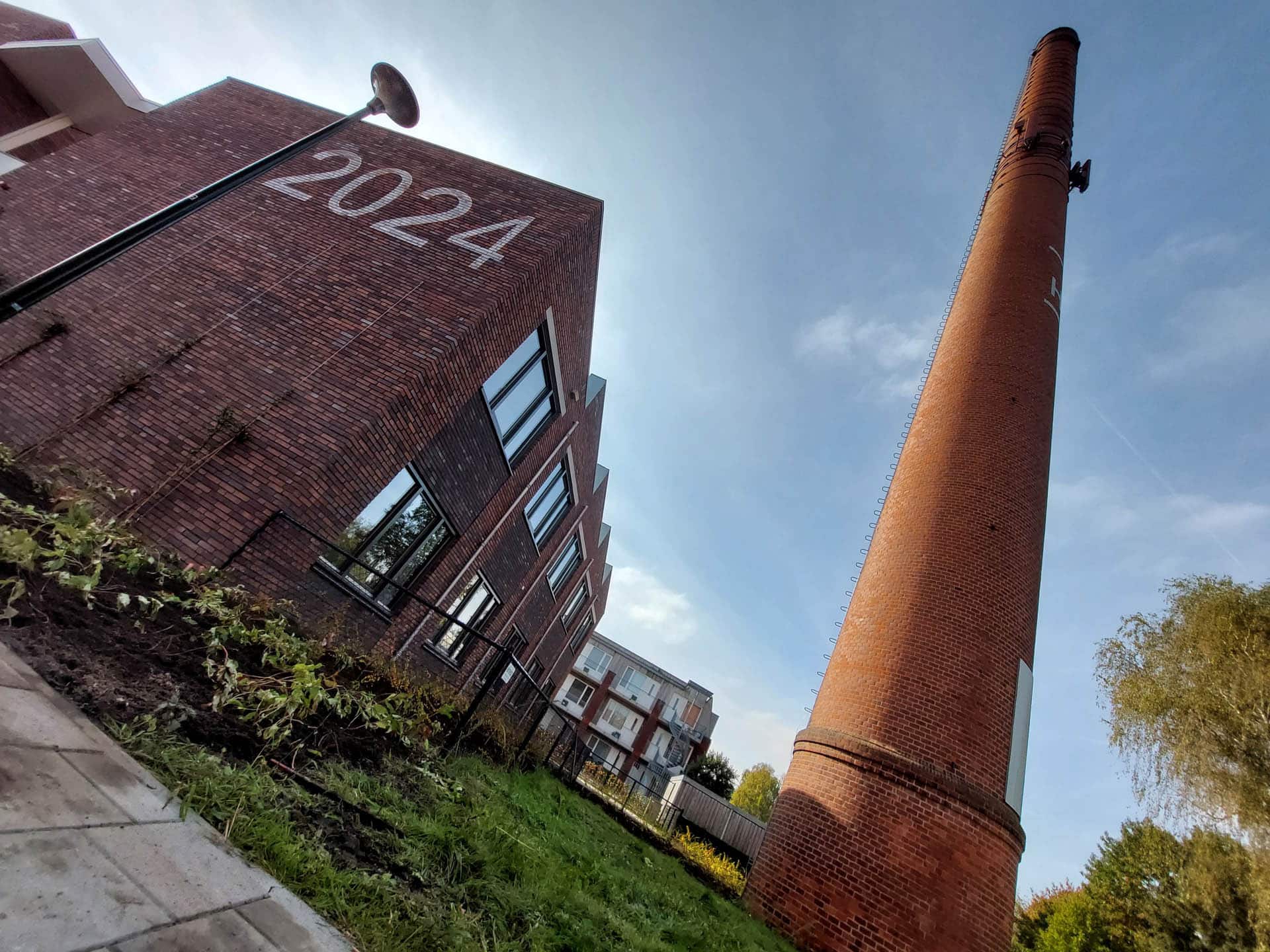 Buitenaanzicht van een rood bakstenen gebouw met het jaartal 2024 op de gevel en een hoge schoorsteen.