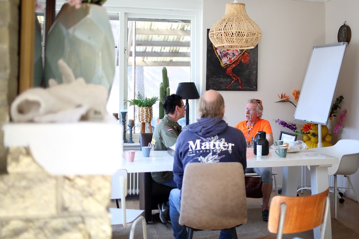 Drie personen in gesprek aan een tafel binnen het Bloezem GGZ Inloophuis Nunspeet, in een huiselijke en kleurrijke ruimte met kunst en planten.