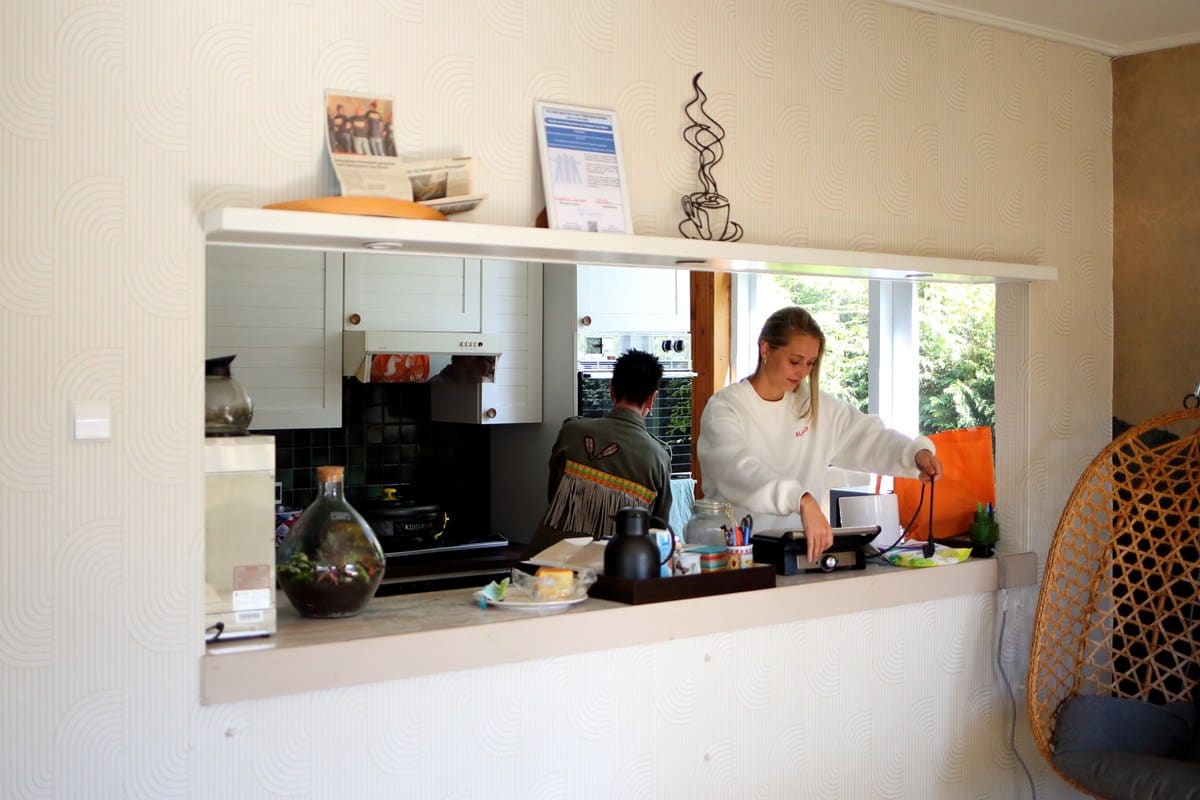 Twee vrijwilligers aan het werk in de keuken van het Bloezem GGZ Inloophuis Nunspeet, met zicht op servies, koffie en een gezellige open keuken.