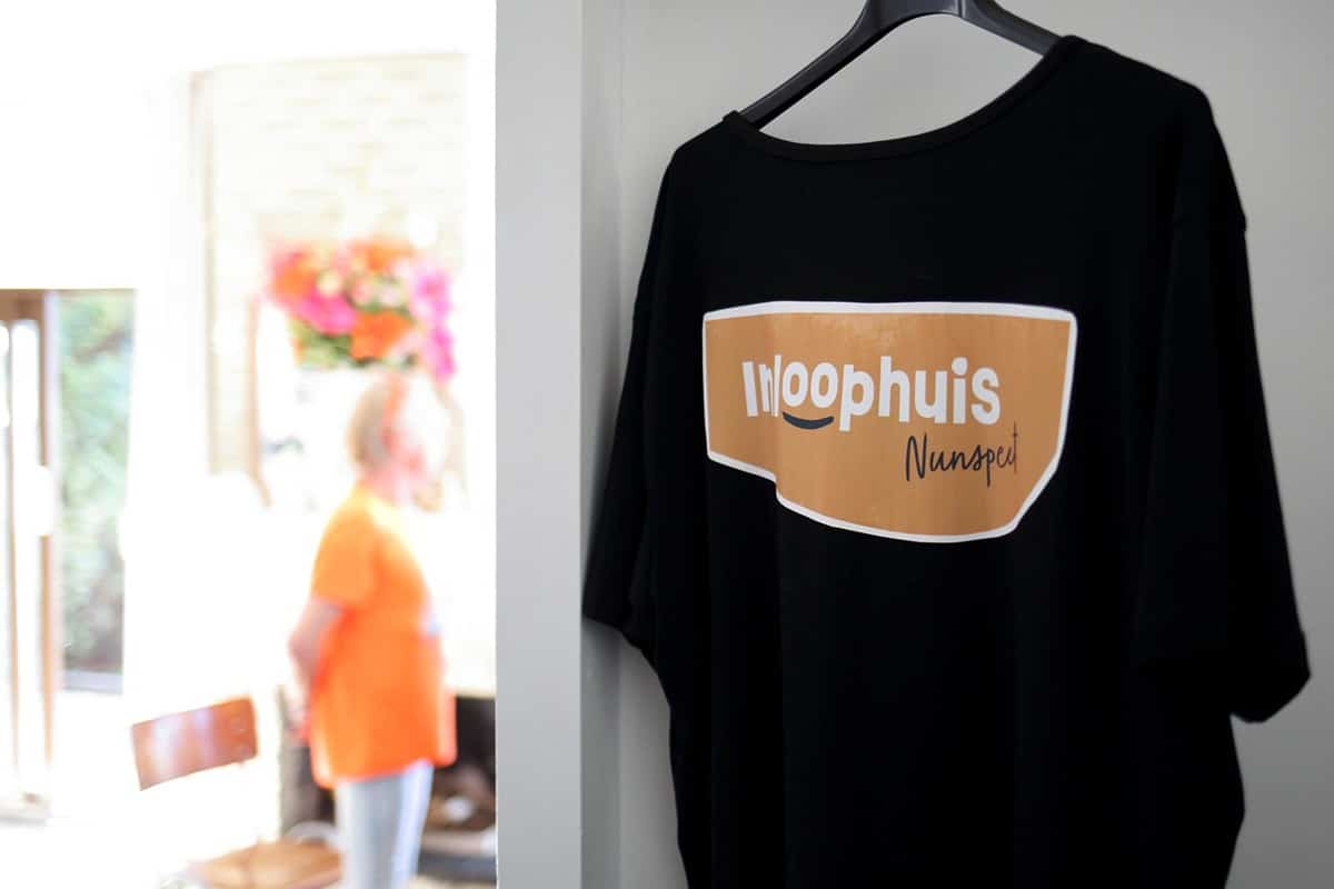 Zwart T-shirt met het logo van Bloezem GGZ Inloophuis Nunspeet aan een hanger, met op de achtergrond een vrijwilliger in oranje shirt in een lichte ruimte.
