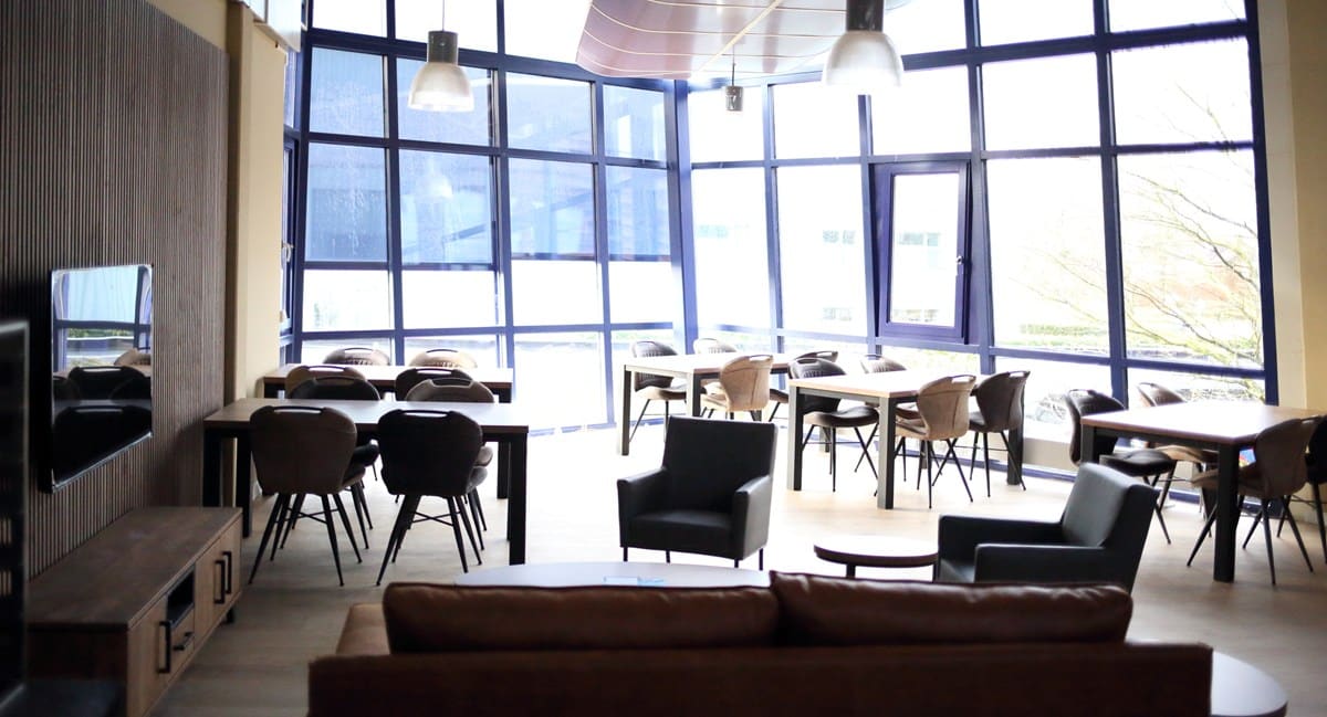 Gemeenschappelijke woonkamer met grote raampartijen, verschillende zitjes en tafels, ingericht met moderne meubels in warme tinten.