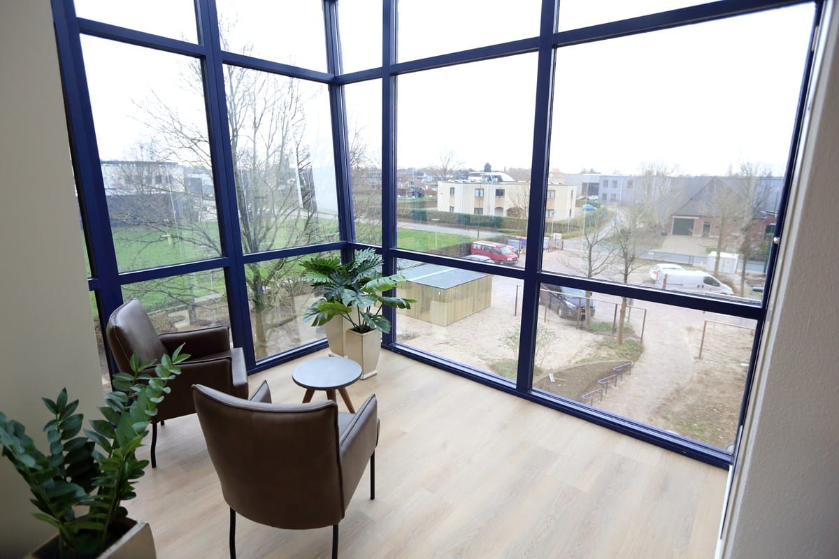 Zithoek in een lichte hoek van het gebouw met twee fauteuils, een klein tafeltje en uitzicht op de woonwijk en het groen buiten.