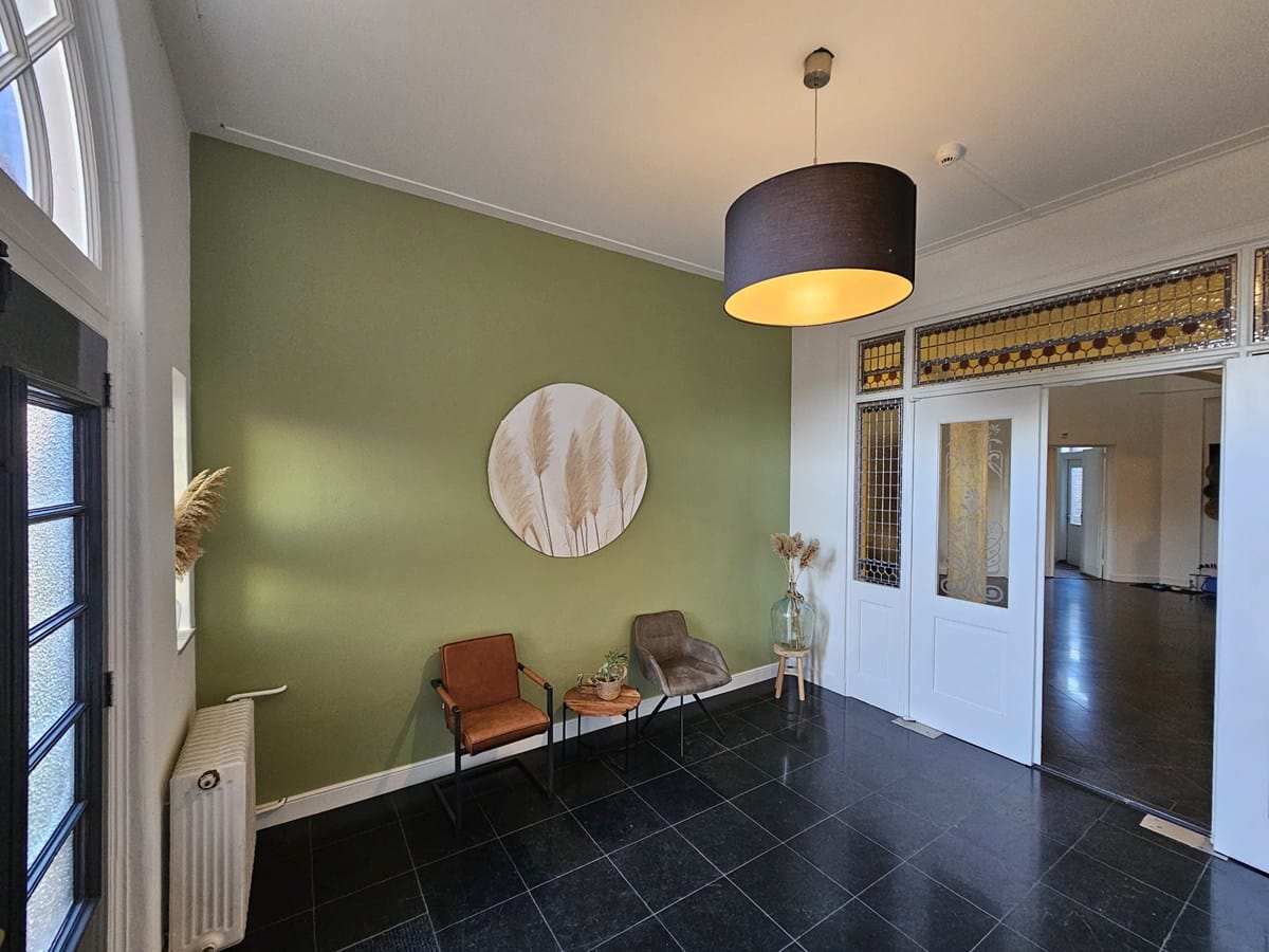 Entree of wachtruimte met een groene muur, moderne verlichting, twee stoelen, een klein tafeltje en een deur met glas-in-loodpanelen.