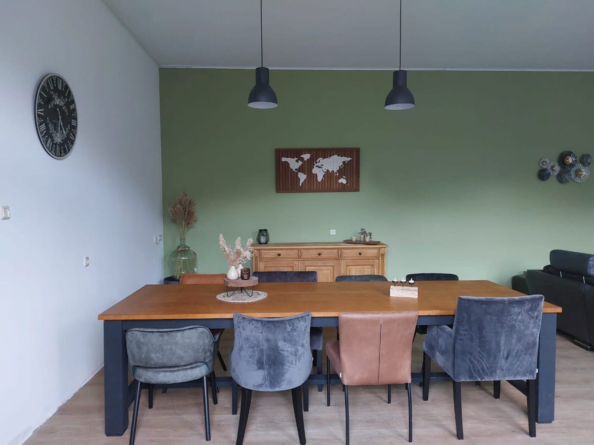 Eetkamer met een grote houten tafel, verschillende stoelen en een groene accentmuur met decoratie, waaronder een wereldkaart en droogbloemen.